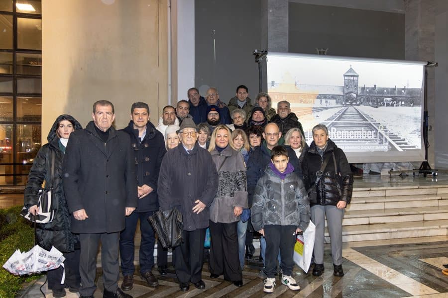 Ανθρώπινη ομάδα μπροστά από φωταγωγημένο Δημοτικό Ωδείο Λάρισας, συμμετέχοντας στην παγκόσμια εκστρα.